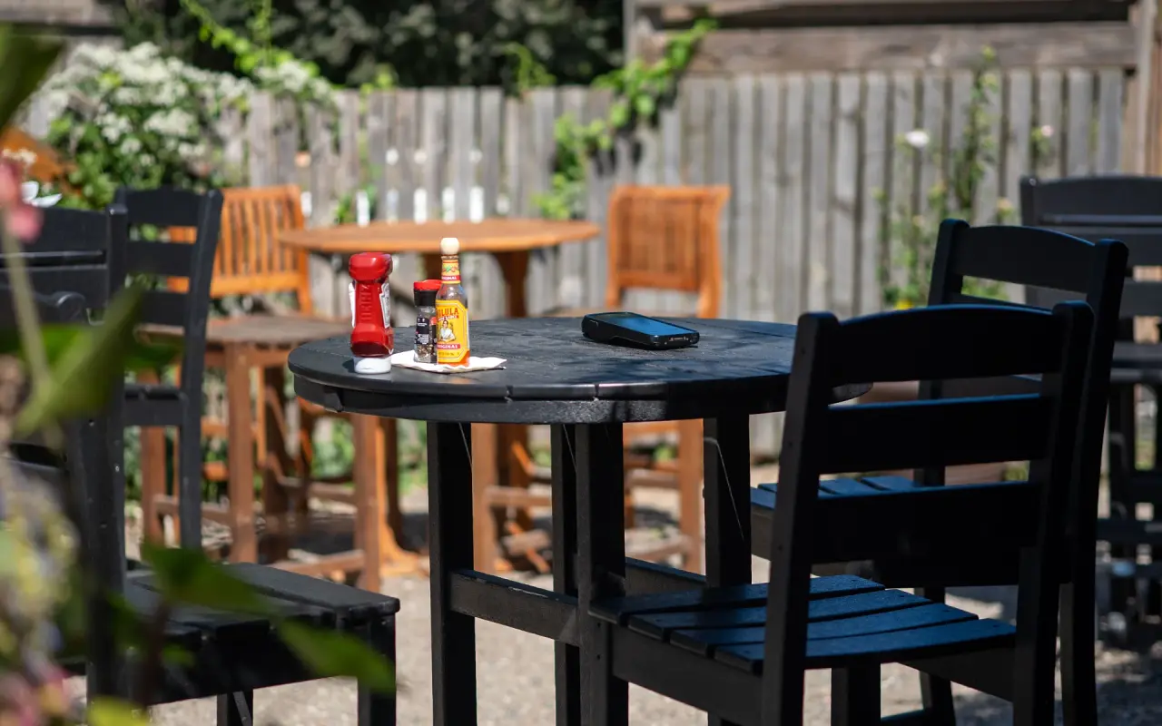 Black outdoor high-top table with ketchup and hot sauce bottles and a phone, wooden chairs behind