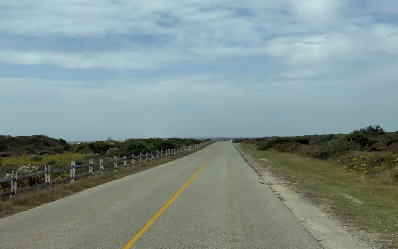 Atlantic Drive behind the dunes at South Beach in Katama, Edgartown: a straight two-lane road with a yellow center line, a weathered split-rail fence on the dune side, goldenrod in bloom among the scrub, a car parked on the grassy shoulder far ahead and a glimpse of the ocean on the left horizon under a pale September sky