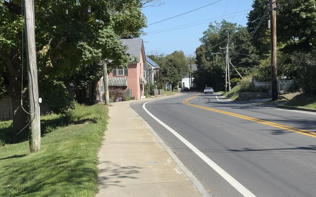 A two-lane road curving into Vineyard Haven under big maples on a sunny September day: a concrete sidewalk on the left, a pink-shingled house with a green striped awning, utility poles and wires, a stone wall and picket fence on the right and a car in the distance