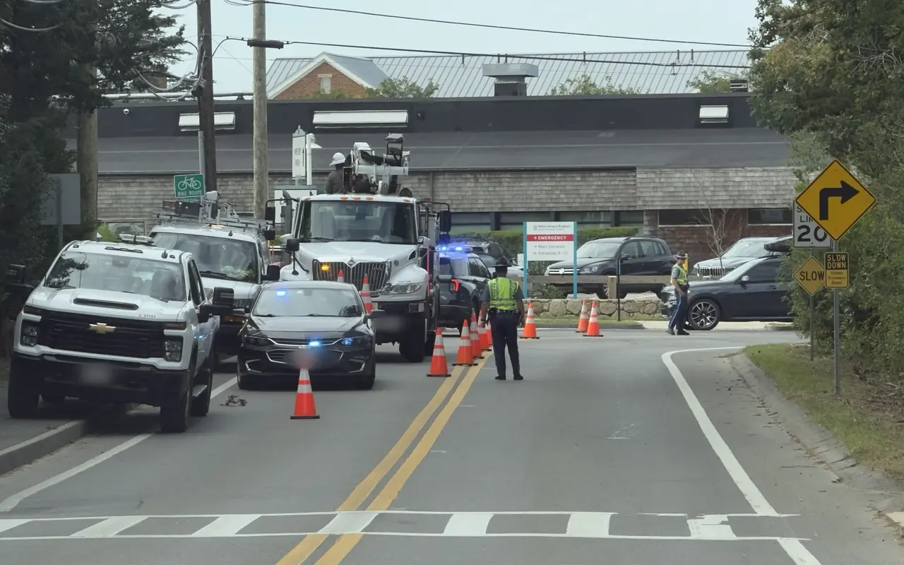 A utility bucket truck and a police detail with traffic cones on Beach Road at the Martha's Vineyard Hospital entrance, with the emergency sign behind