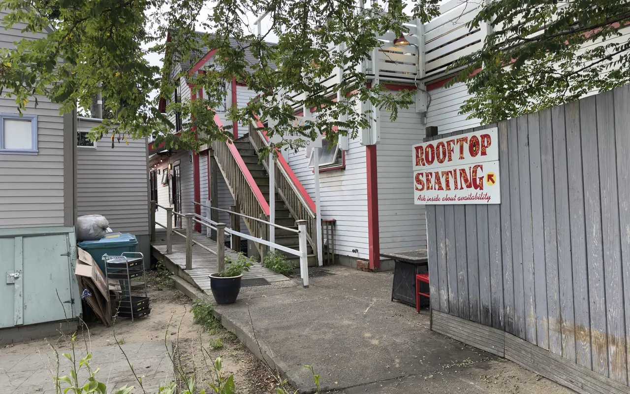 A hand-painted Rooftop Seating sign with an arrow on a grey fence in an alley in Oak Bluffs, Ask inside about availability, a white building with red trim and an outside stair up to a roof deck behind it