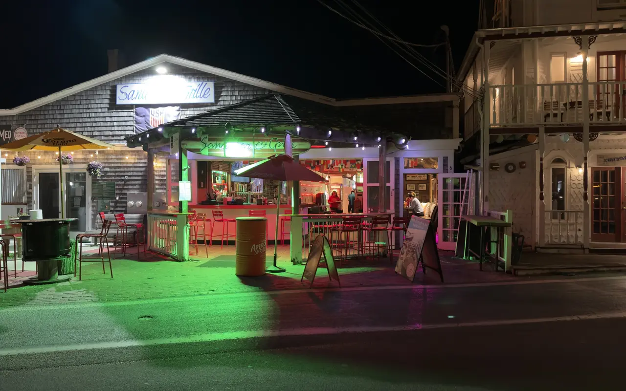 The Sand Bar and Grille on Oak Bluffs harbor at night: open-air bar under string lights, red stools, an Aperol umbrella and green and red floodlight on the pavers