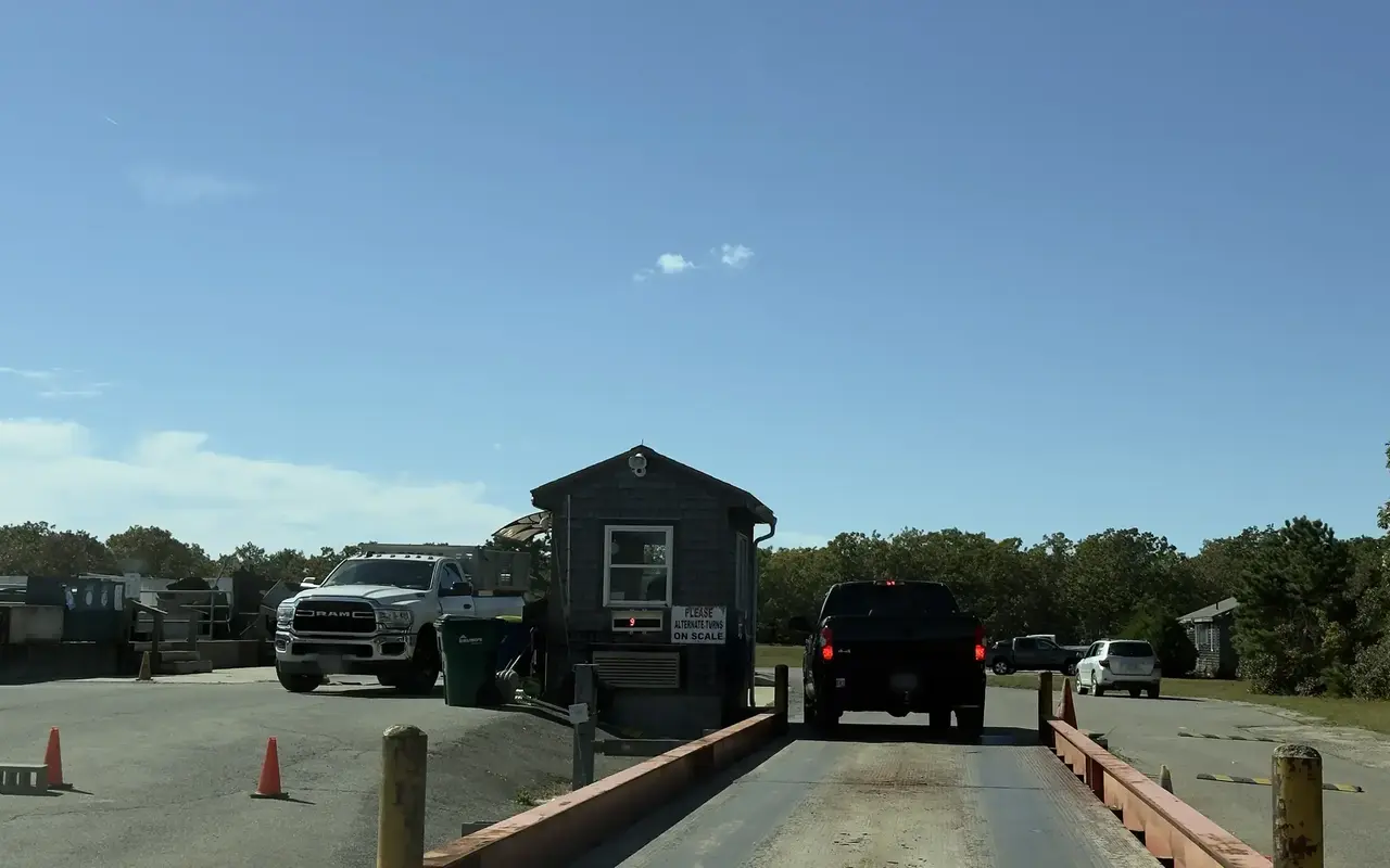 Shingled scale house at the Refuse District transfer station in Edgartown, a dark pickup on the scale and a white pickup parked beside the hut