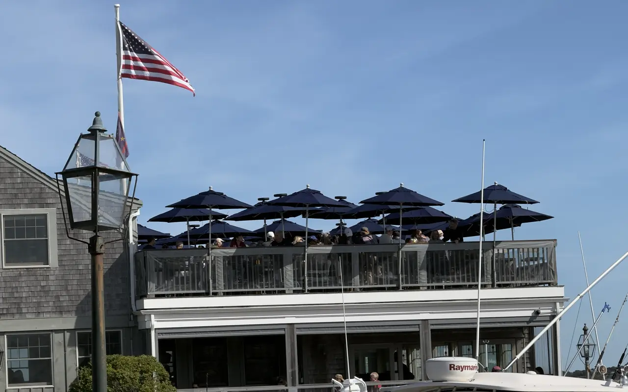 A rooftop restaurant deck crowded with diners under rows of navy umbrellas, an American flag above and a sportfishing boat in the foreground