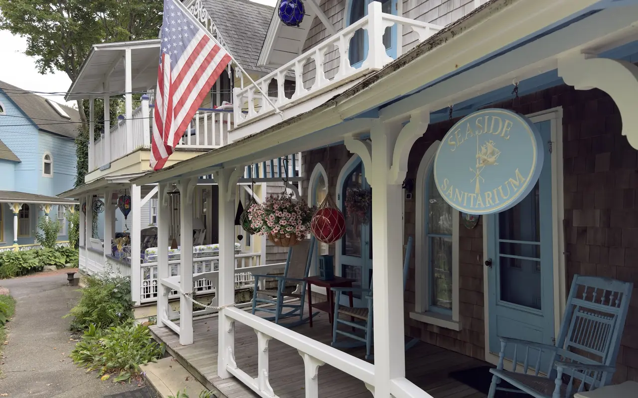 A round blue sign reading Seaside Sanitarium with a ship on it, hung on the porch of a grey-shingled cottage with white brackets, blue trim, blue rocking chairs, a hanging basket of pink petunias, glass fishing floats and an American flag, in the Oak Bluffs Campground