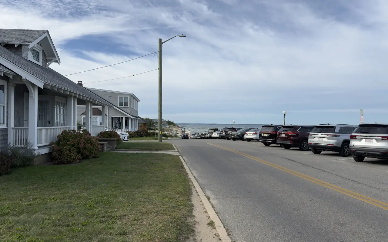 A residential street in Oak Bluffs sloping toward the water: grey-shingled cottages with white porches and hydrangeas on the left, a row of SUVs angle-parked on the right, and Nantucket Sound and a pier at the end under a high cloudy sky