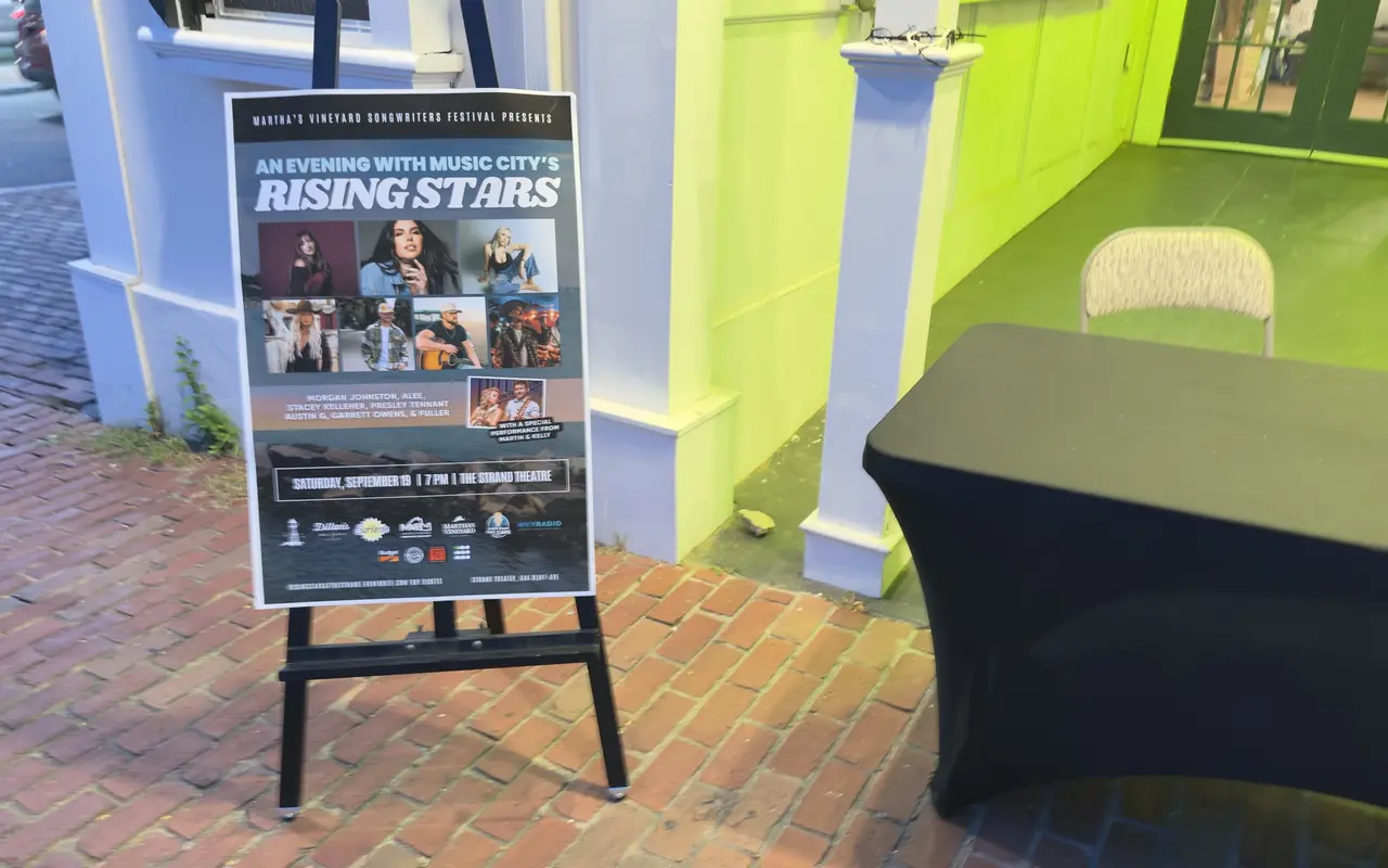 An easel outside the Strand Theatre on Oak Bluffs Avenue with the Rising Stars poster for the Martha's Vineyard Songwriters Festival: seven performer photos, the date Saturday September 19 at 7 PM, and the sponsor logos along the bottom, a black-draped table beside it on the brick sidewalk