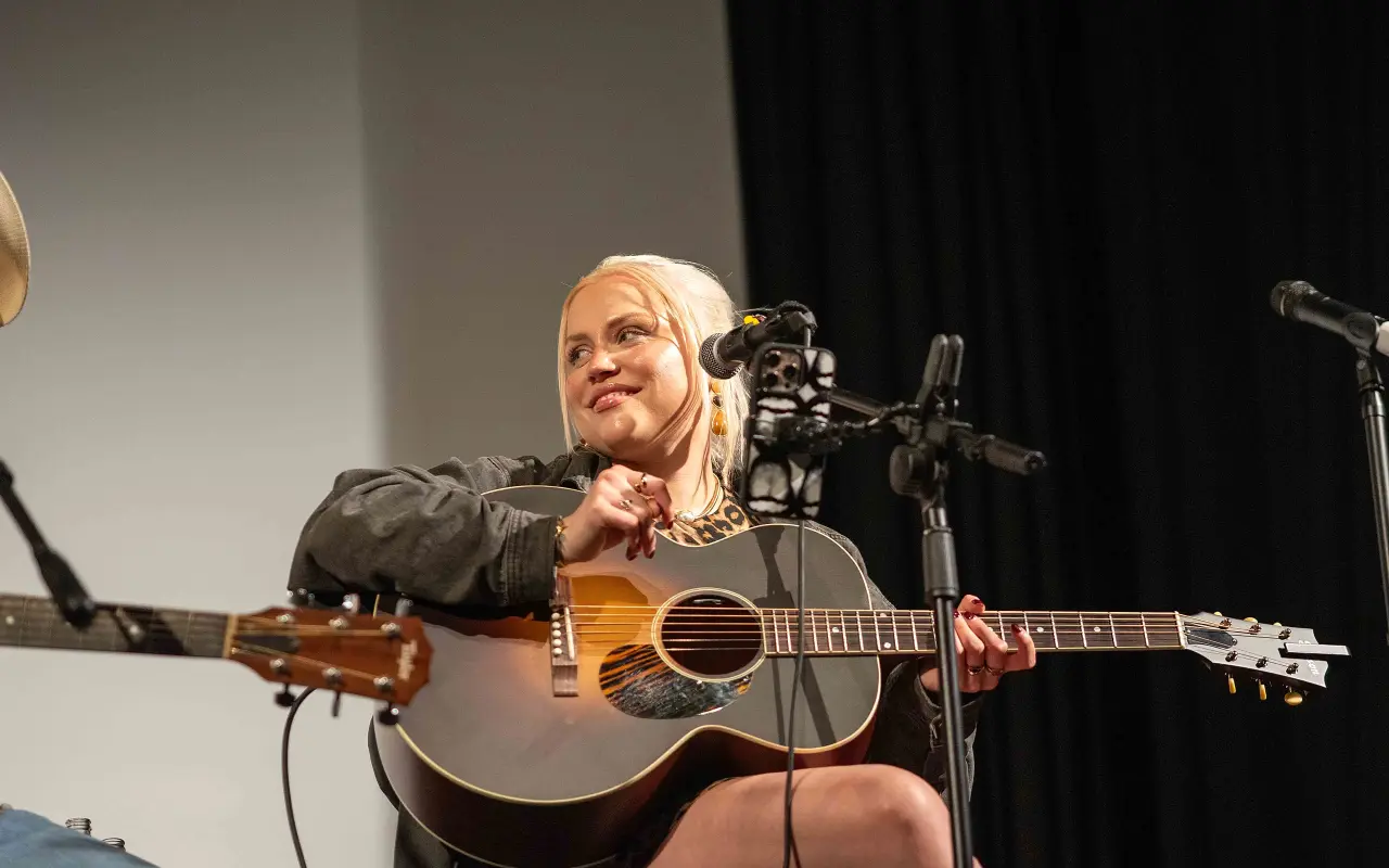 Stacey Kelleher resting her arm on an acoustic guitar and smiling on the Strand stage