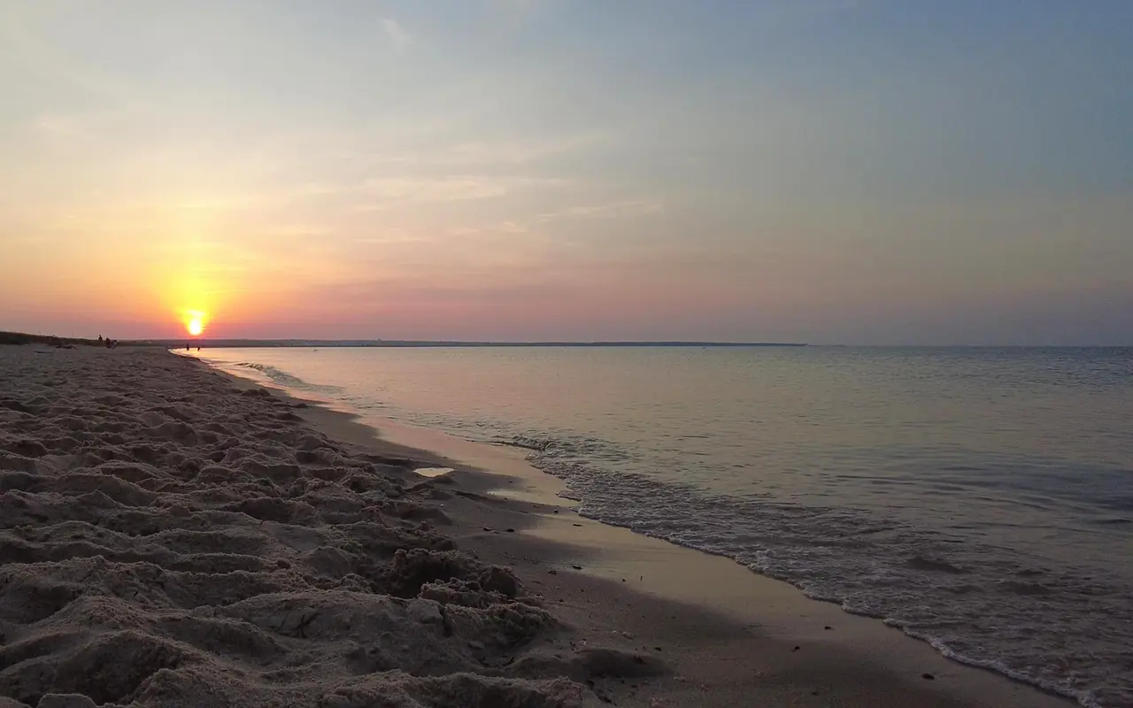 The sun setting low over the water at the far end of Joseph Sylvia State Beach, footprints in the sand and calm water along the shore