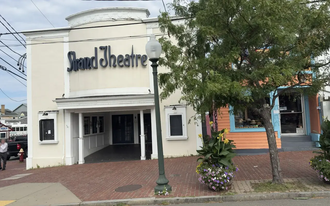 The cream facade of the Strand Theatre on Oak Bluffs Avenue with its dark script sign, a white columned entrance and blank poster cases, a green lamppost, a tree and flower barrels on the brick sidewalk, an orange hat and sunglasses shop next door and bike rentals across the street
