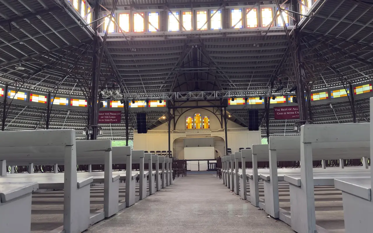 Inside the Tabernacle in Oak Bluffs: rows of white wooden benches, the iron roof trusses, stained glass along the clerestory and the stage with three arched windows at the end