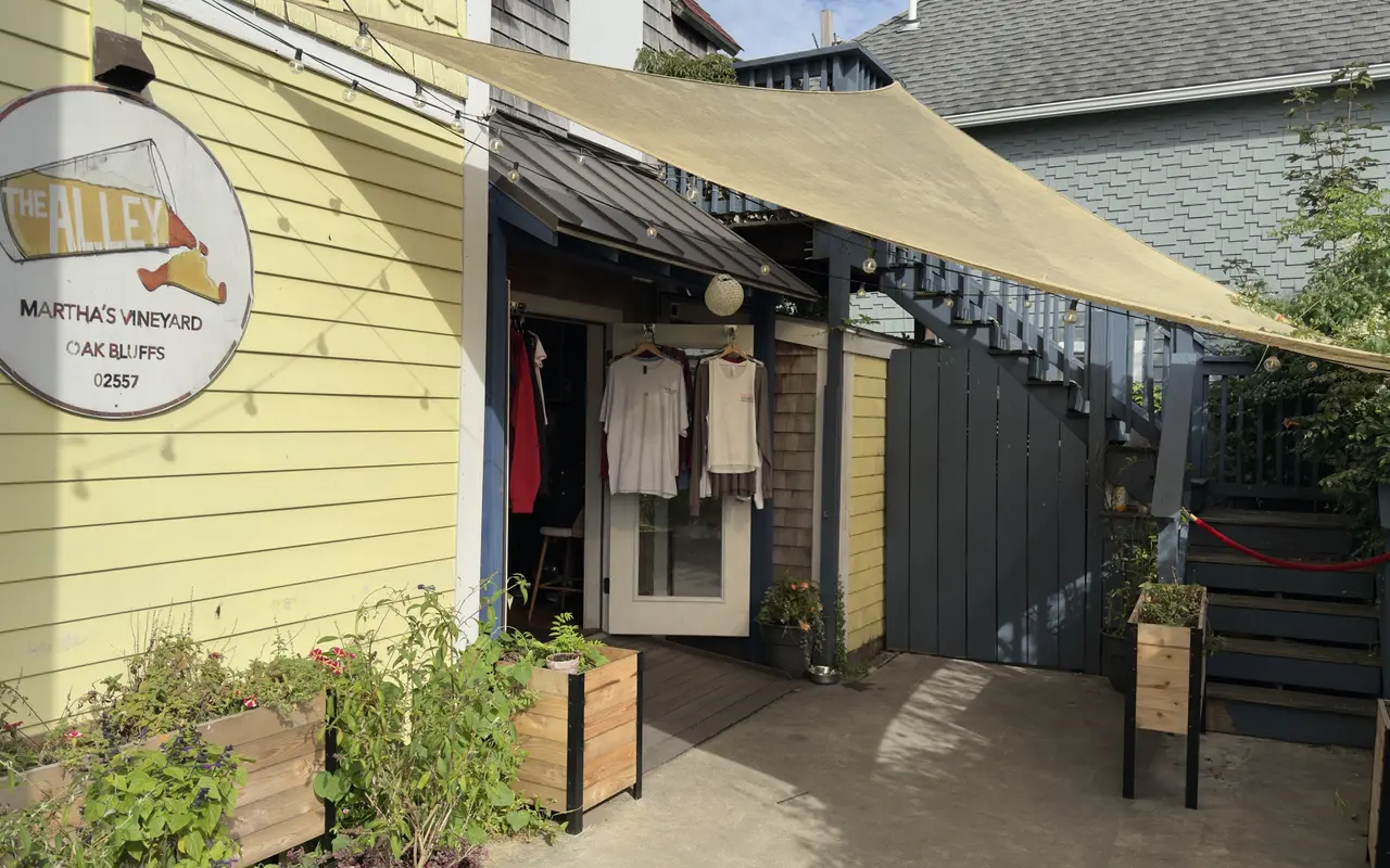 The entrance to The Alley bar in Oak Bluffs: a round sign on a yellow clapboard wall, a shade sail over a concrete walk, T-shirts and sweatshirts hanging in the open door, planter boxes with herbs and flowers, a grey stair and a red rope beside it