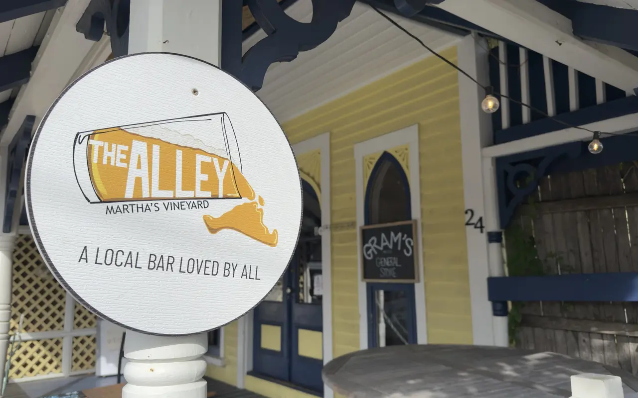 A round white sign for The Alley, Martha's Vineyard, a local bar loved by all, with a tipped beer glass pouring out the shape of the island, on a white porch post in Oak Bluffs, a yellow building with a Gram's General Store chalkboard and the number 24 behind it