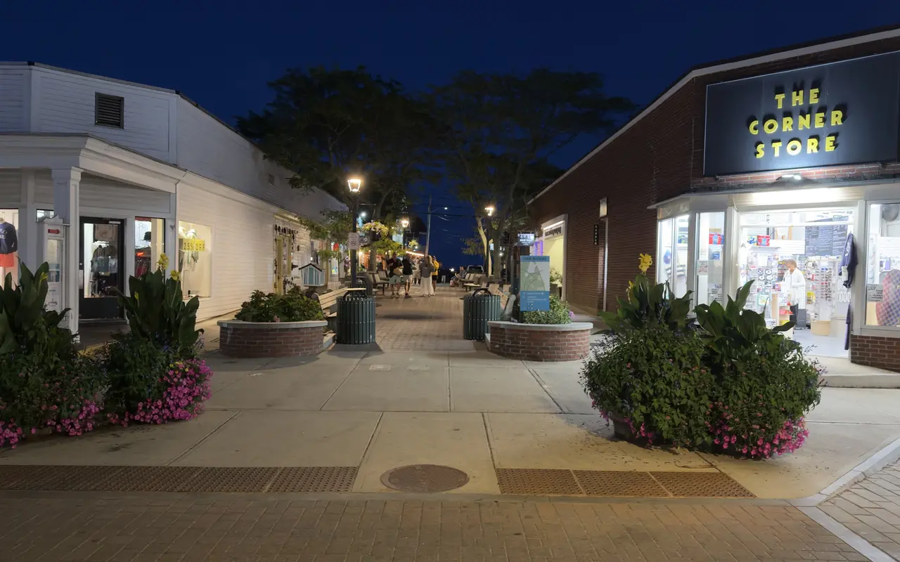 The Corner Store and the Kennebec Avenue plaza in Oak Bluffs at dusk, with brick planters, a lamp post and shoppers under the trees