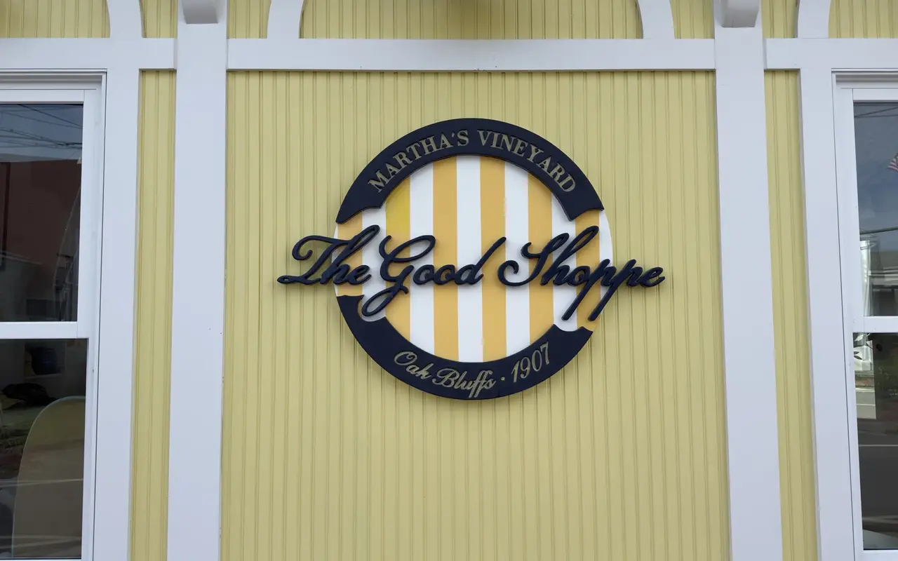 Round navy and yellow striped sign reading The Good Shoppe on a yellow clapboard wall