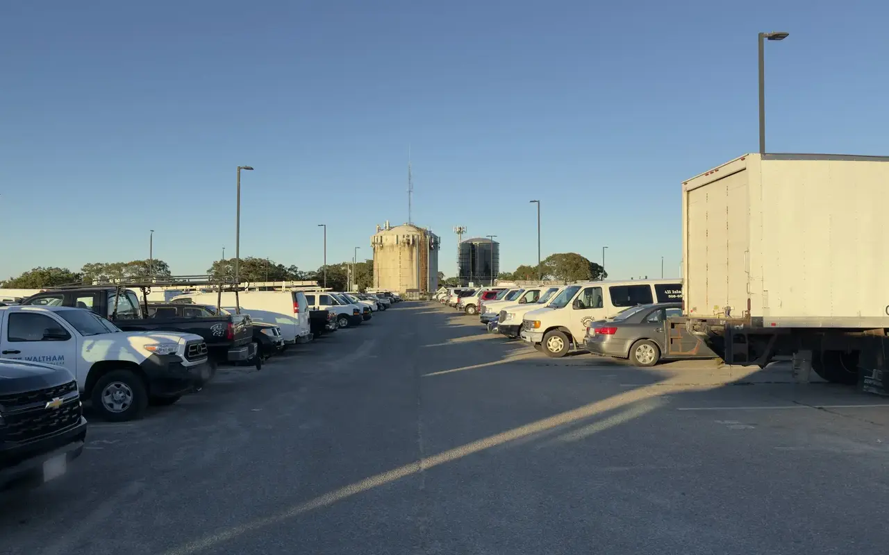 A wide paved parking lot at the Tisbury Park and Ride in Vineyard Haven with rows of white work vans, pickups and a box truck, two tall round storage tanks at the far end under a clear sky