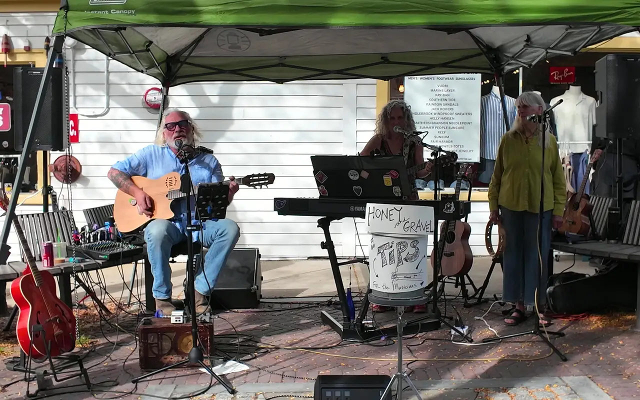 Three musicians with guitar, keyboard and vocals playing under a green pop-up tent on a brick sidewalk, a tip bucket in front