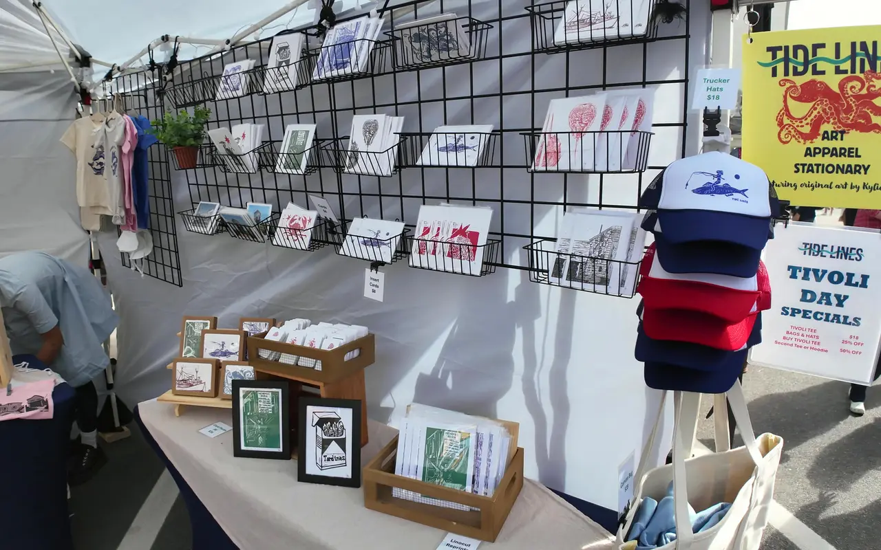 A vendor booth with block-print cards on a wire grid, framed prints, stacked trucker hats and a yellow Tide Lines sign