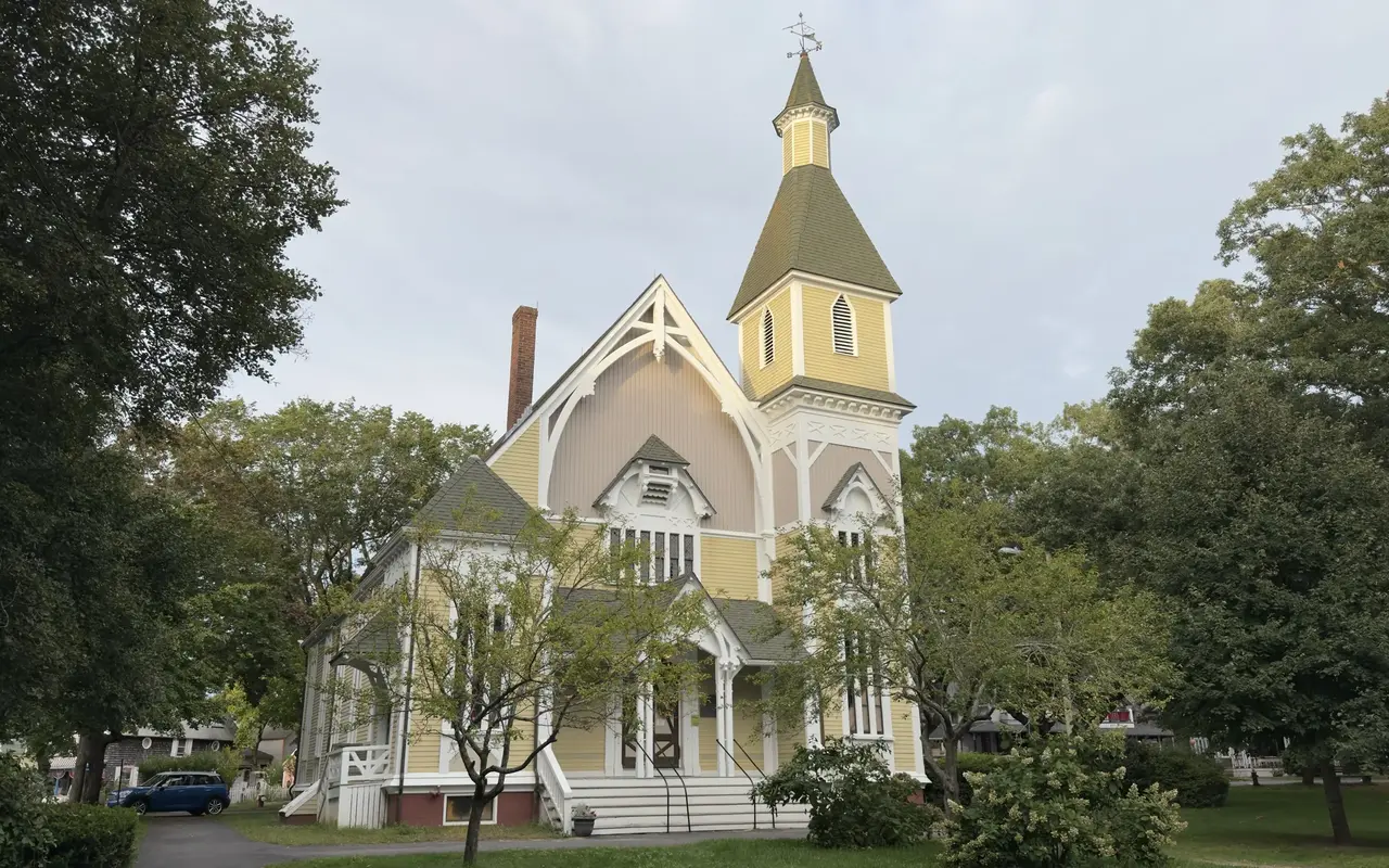 Trinity Methodist Church in the Oak Bluffs Campground, a yellow Carpenter Gothic church with white trim, a pointed steeple and a weathervane, framed by oak trees
