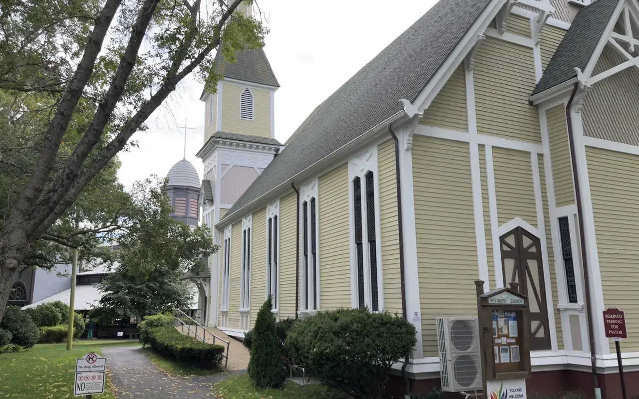 The long side of Trinity Methodist Church in Oak Bluffs, yellow clapboard with white Gothic trim and tall windows, the Tabernacle's cupola behind it, a No Dogs Allowed sign on the path