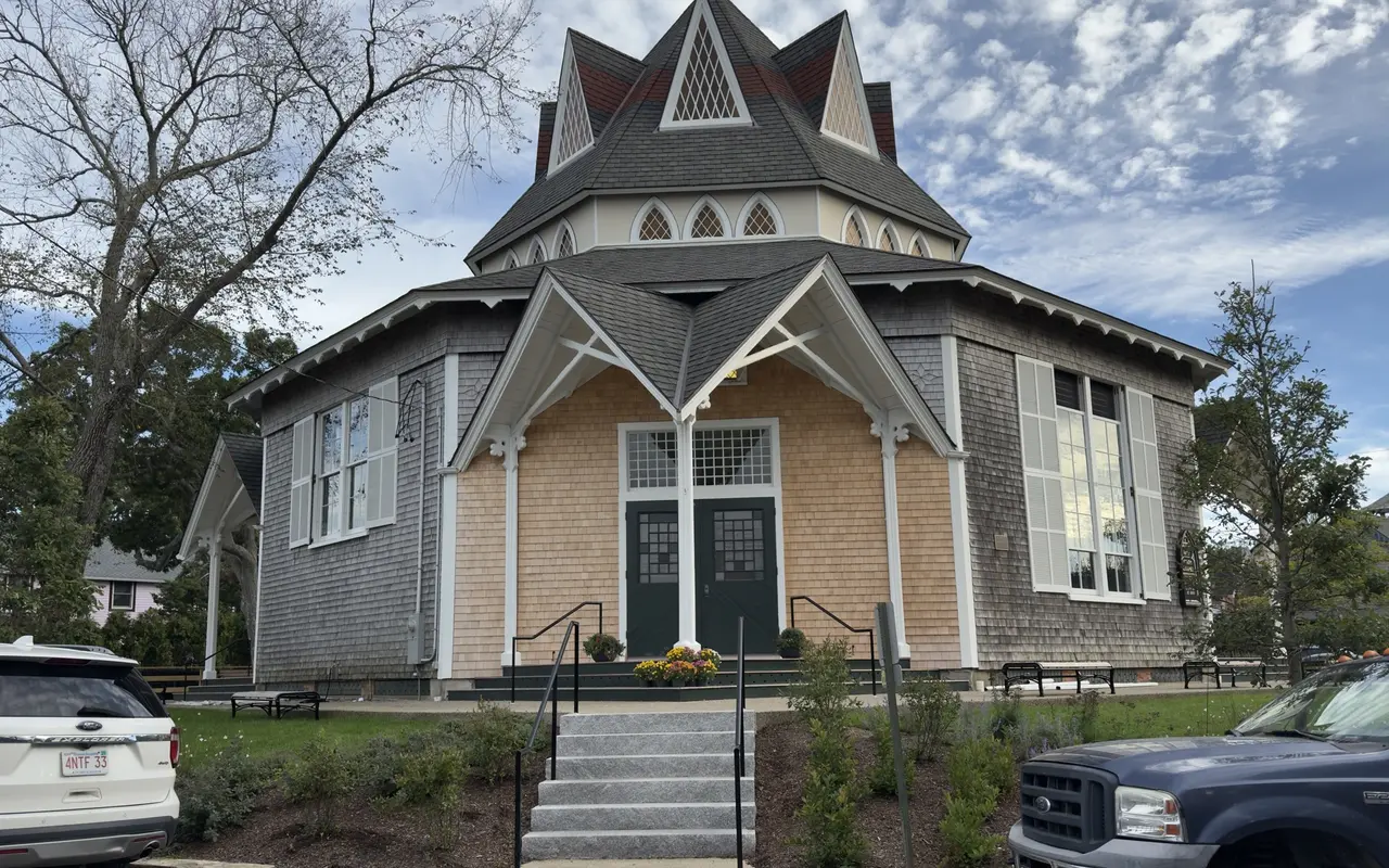 Union Chapel in Oak Bluffs seen from the front: an octagonal shingled chapel with a tiered roof, pointed dormers with lattice windows, white brackets over the entrance, green double doors, granite steps and marigolds by the door