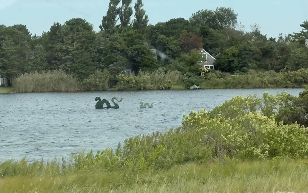 Two green sea serpent sculptures, one big and one small, floating on Farm Pond in Oak Bluffs with a house and trees on the far shore