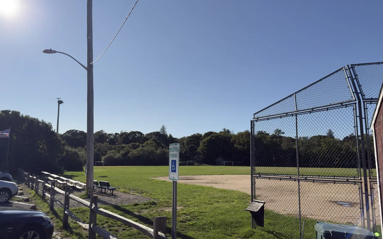 Veterans Memorial Park in Vineyard Haven: wooden bleachers along a split-rail fence, the ball field and backstop, an American flag and an osprey nest pole in the sun