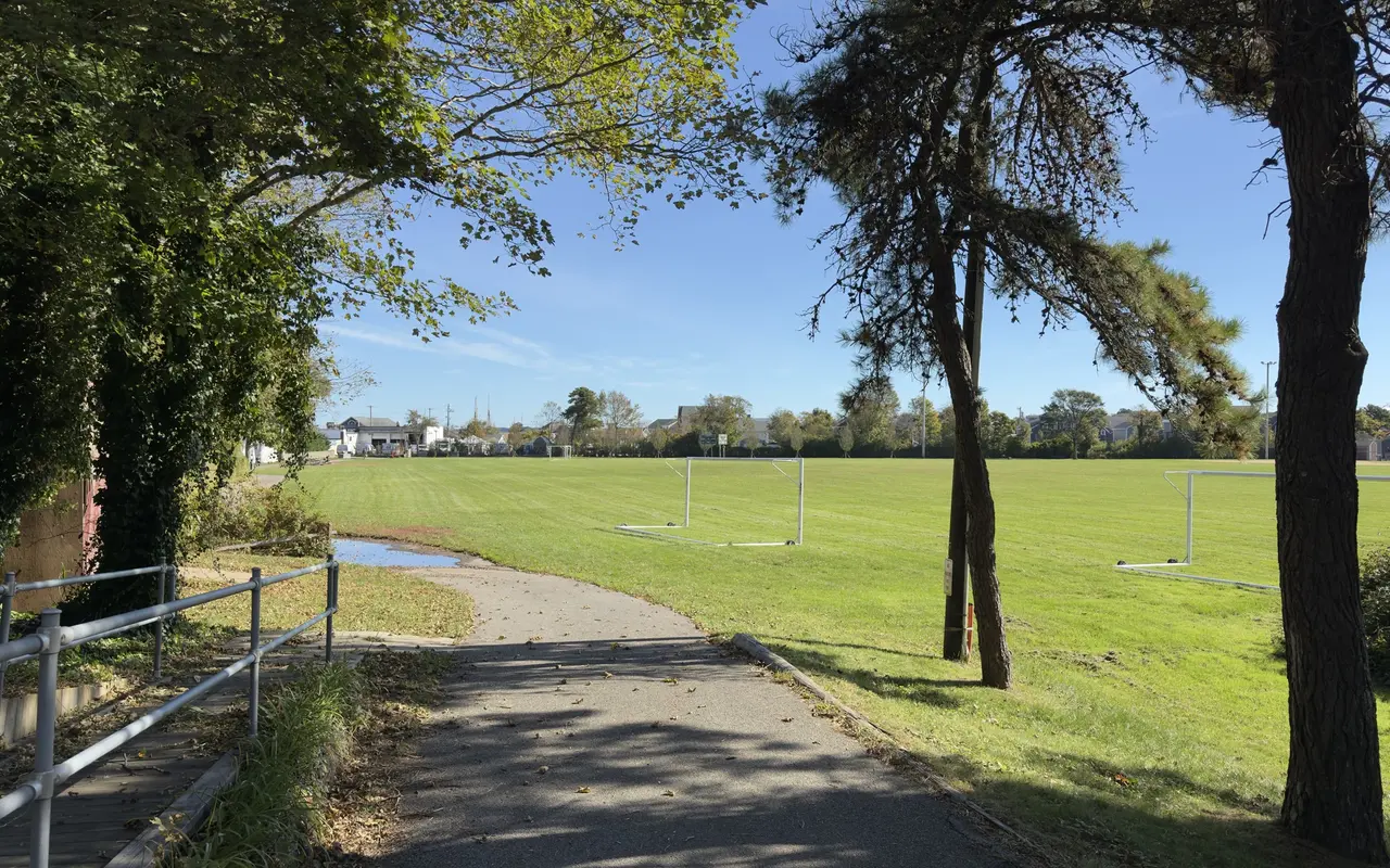 A paved path under trees beside a wide green field with soccer goals at Veterans Park in Vineyard Haven
