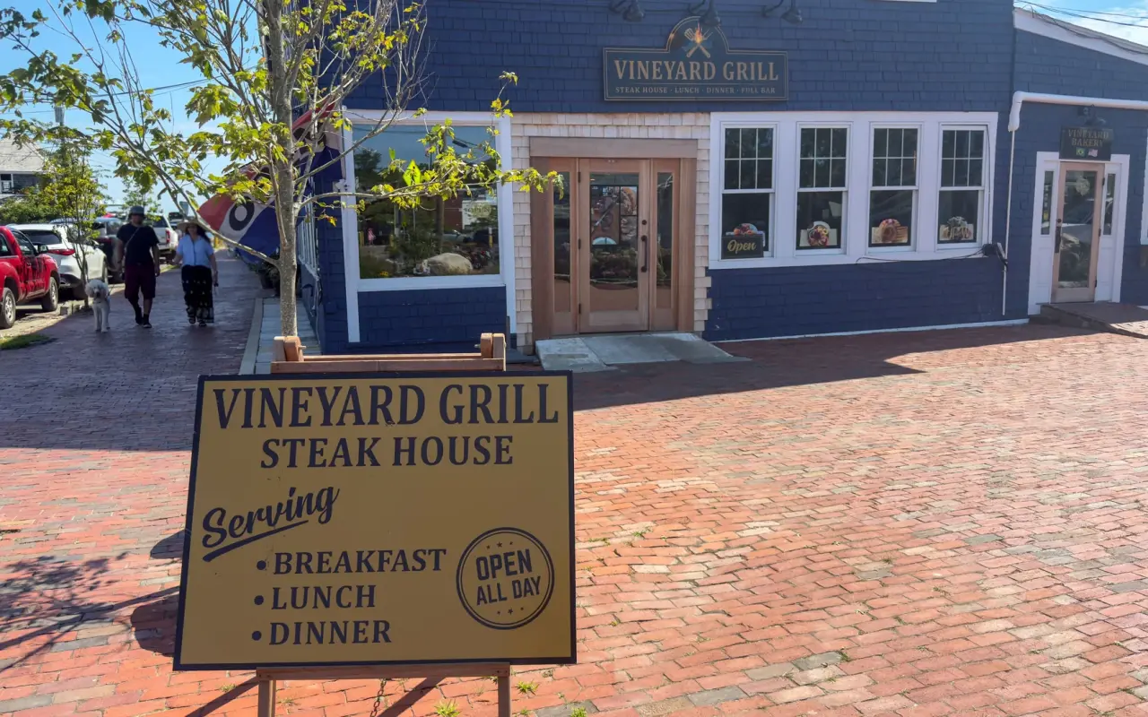 A sandwich board sign on a brick sidewalk reading Vineyard Grill Steak House, breakfast lunch dinner, in front of a navy storefront