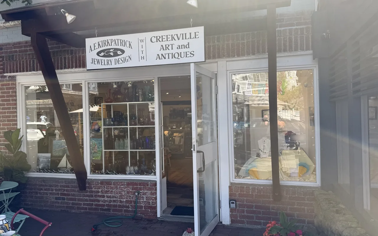 A brick shopfront with a white sign reading A.E. Kirkpatrick Jewelry Design with Creekville Art and Antiques, an open door, glass vases and bottles on white shelves in the window and jewelry on display