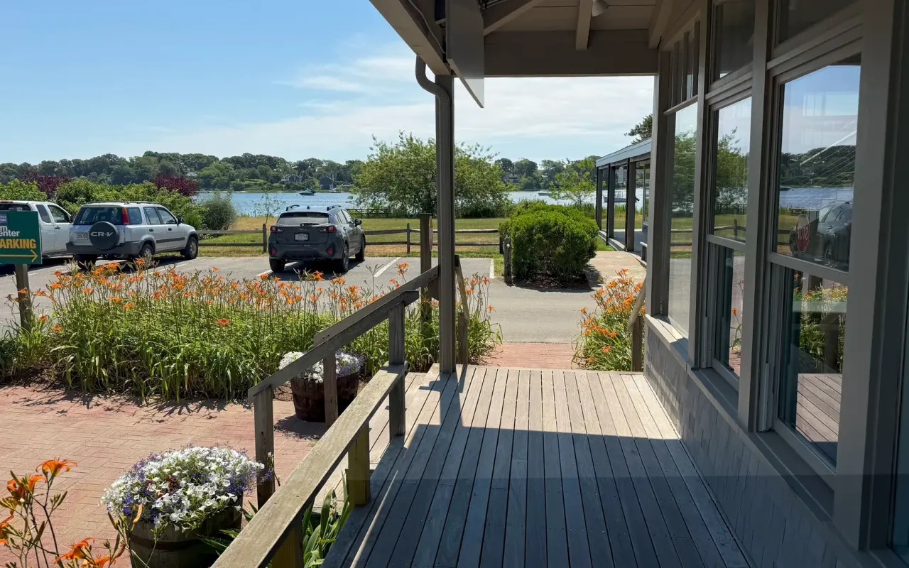 Wooden deck at Tisbury Marketplace in Vineyard Haven with daylilies, a parking lot, a Film Center parking sign and Lagoon Pond beyond