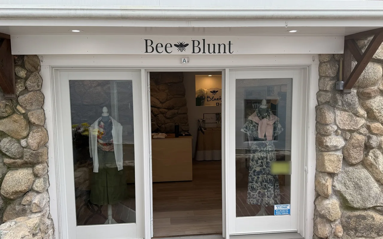 A white shopfront set into a fieldstone wall with the sign Bee Blunt and a bee logo, glass doors open, mannequins in the windows wearing a white cardigan and a blue toile dress with a pink sweater