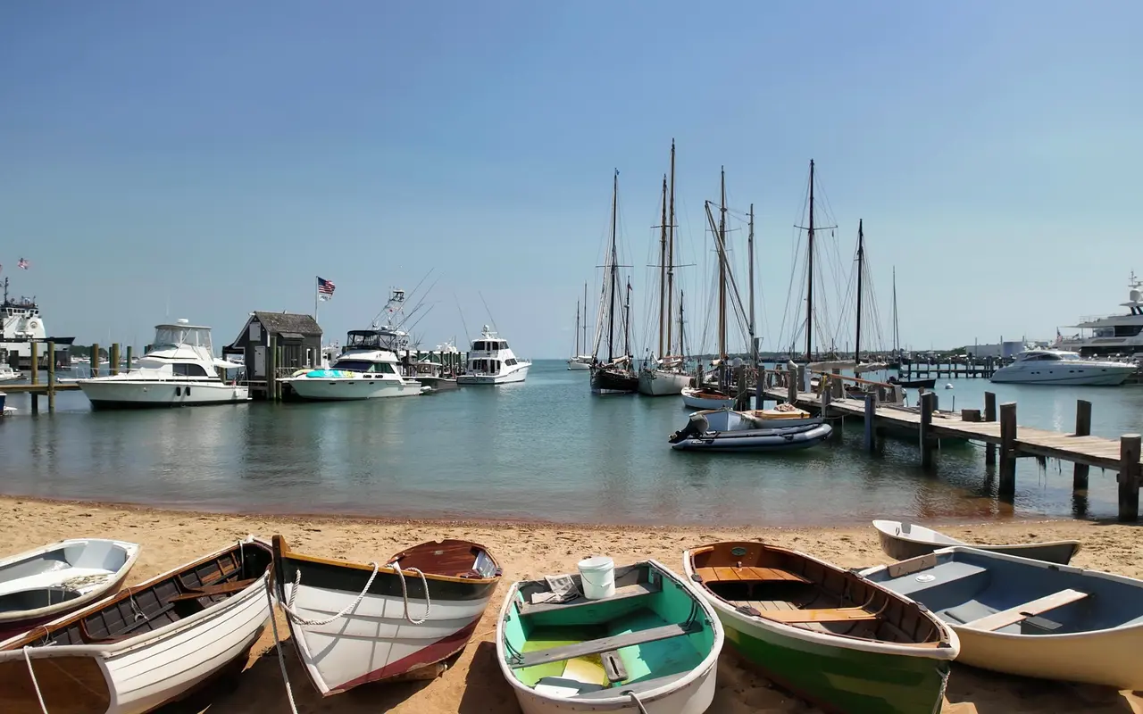 A row of small wooden rowing dinghies on a sandy harbor beach, schooner masts, motorboats and docks in the background