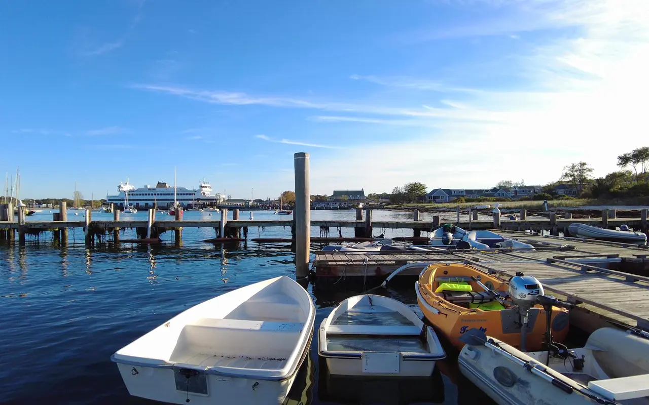 White, orange and gray dinghies tied at a wooden float in Vineyard Haven Harbor, a large white ferry docked across the water under a blue sky