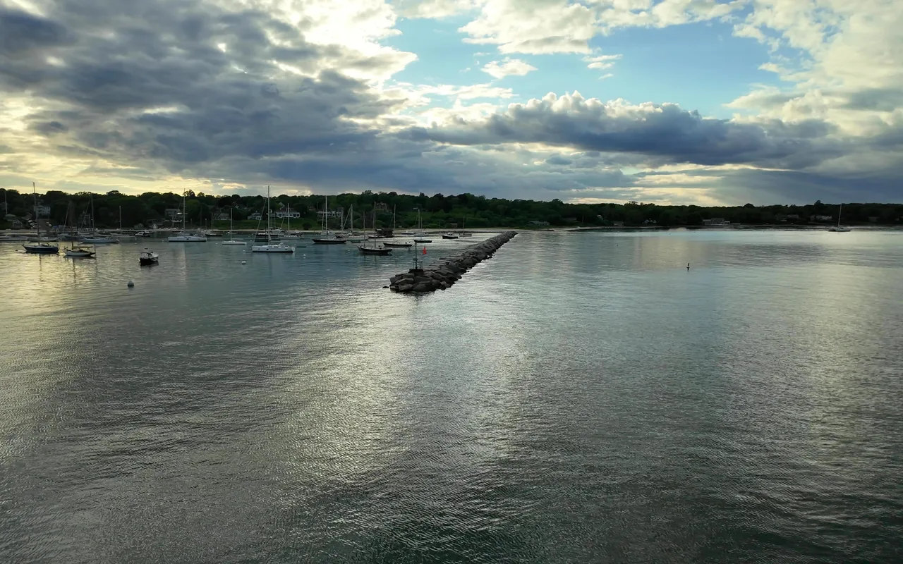View from a departing ferry over Vineyard Haven harbor with a stone breakwater, moored sailboats and a cloudy sky
