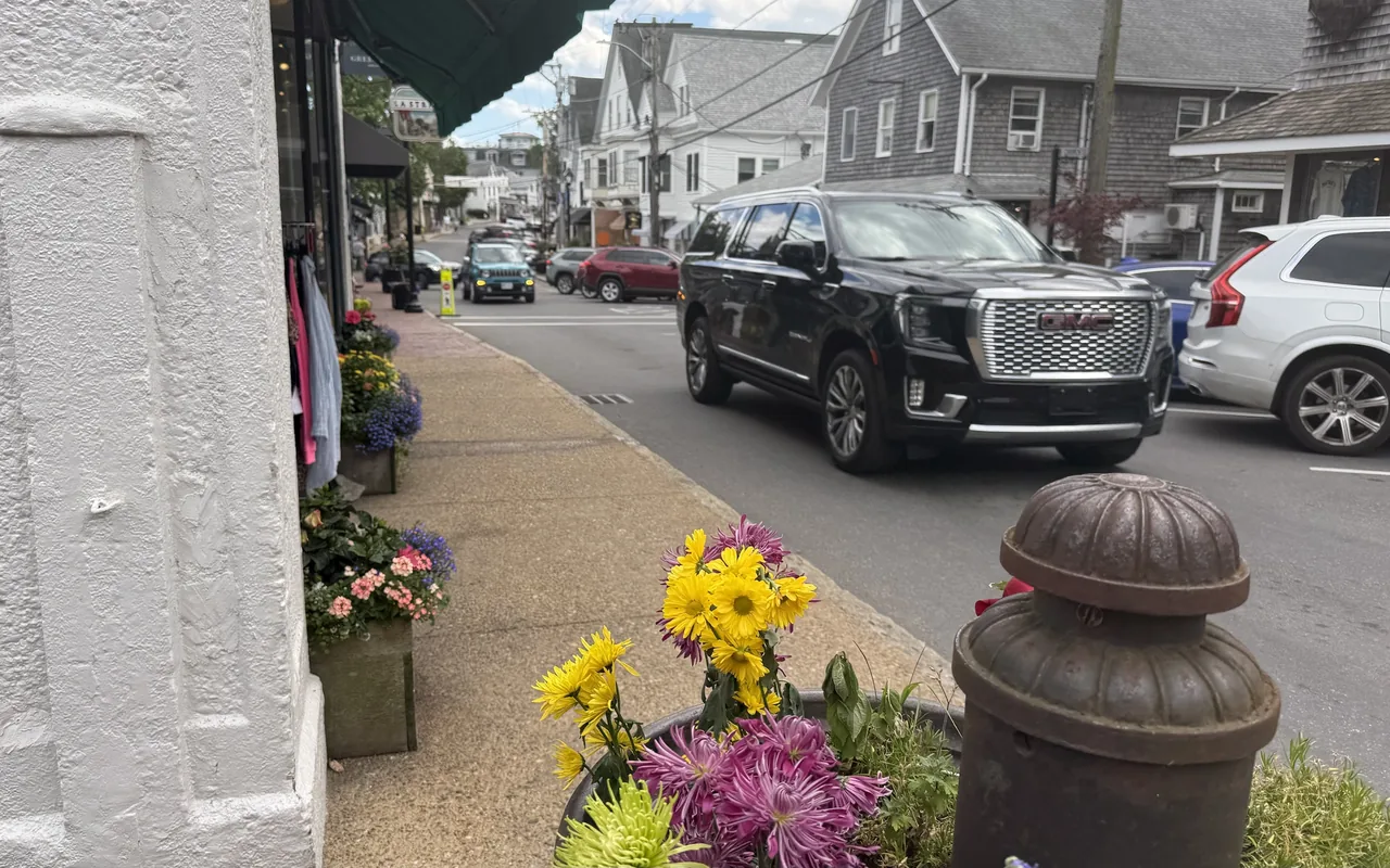 A Vineyard Haven Main Street sidewalk with flower boxes and planters of mums beside shopfronts, a black SUV and other cars on the street and gray-shingled buildings beyond