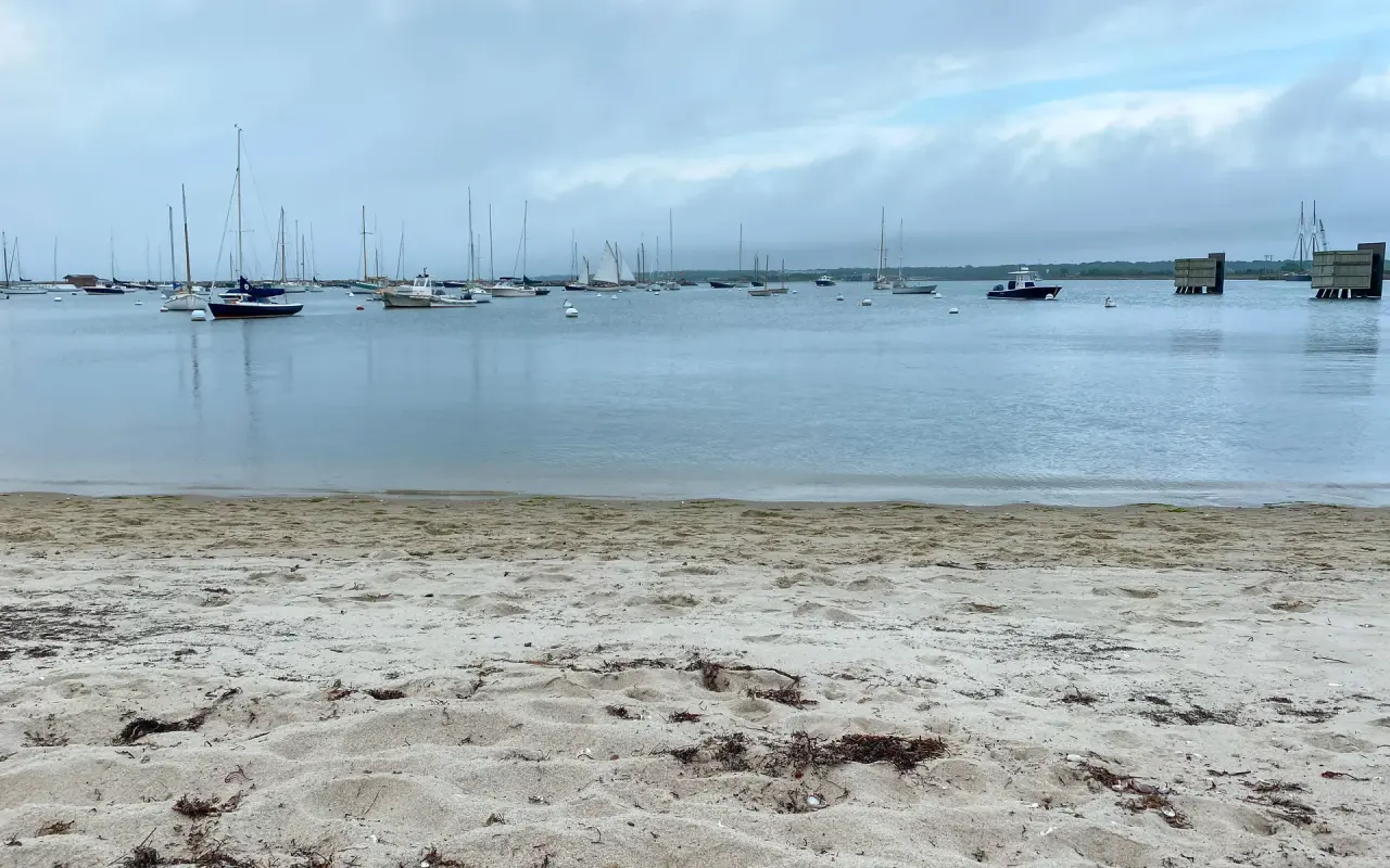 Vineyard Haven harbor under grey cloud with a few moored boats and an empty sandy beach in the foreground