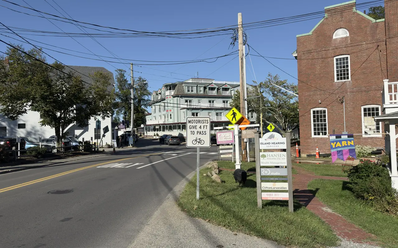 State Road in Vineyard Haven looking toward the Mansion House hotel on Main Street, with a Motorists Give 4 Ft to Pass bicycle sign, the brick Educomp building and a stack of business signs on the right and Brickman's on the left