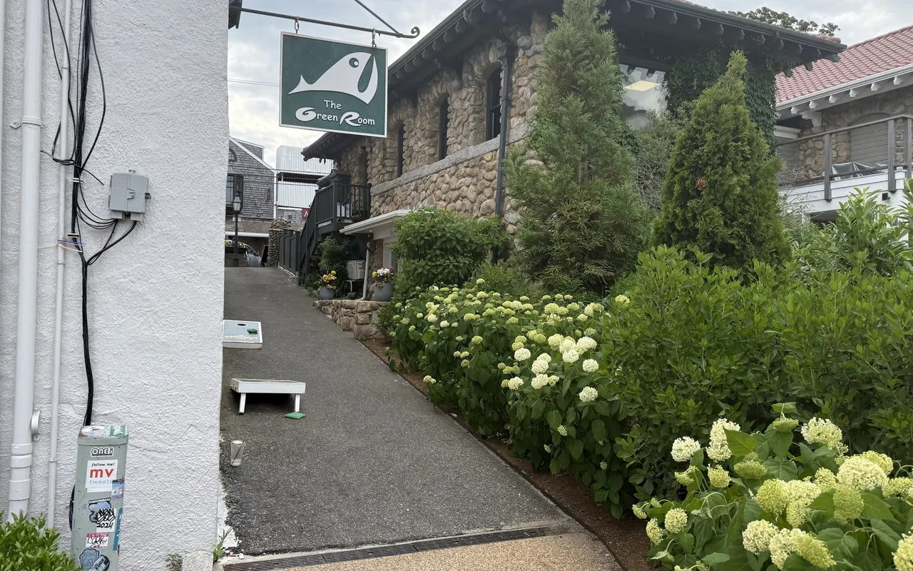 A hanging green sign with a white fish reading The Green Room over a narrow paved path beside a fieldstone building, white hydrangeas and evergreens along the wall and a cornhole board on the path