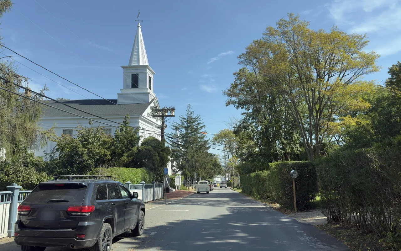 A quiet tree-lined street with a white church building and steeple on the left behind a picket fence, a parked car in front