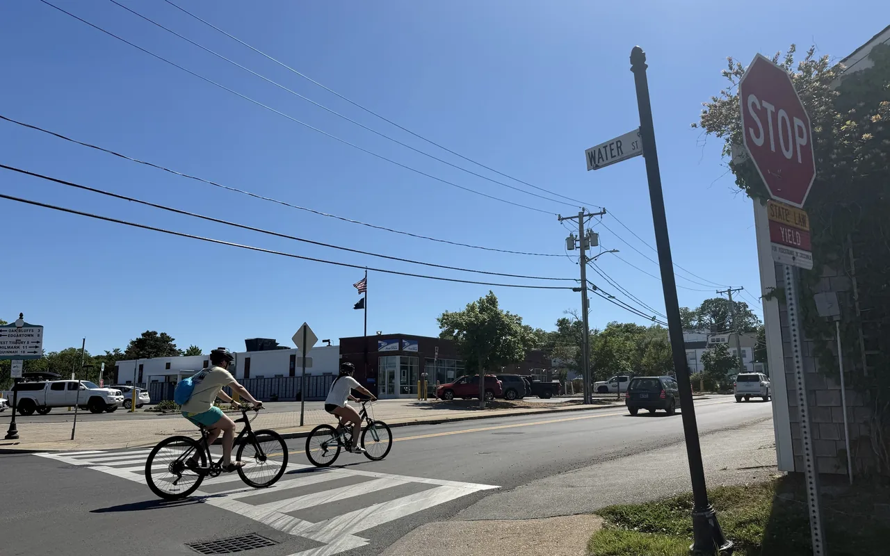 Two cyclists in helmets crossing a crosswalk at a Water Street corner in Vineyard Haven, a stop sign and Water St street sign on the right, the brick and white post office building behind and a white mileage sign listing the island towns on the left