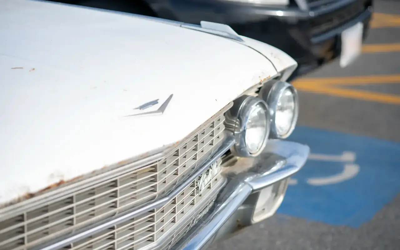 Close-up of the front of a vintage white car with a chrome grille and twin round headlights, a blue accessible parking symbol on the pavement