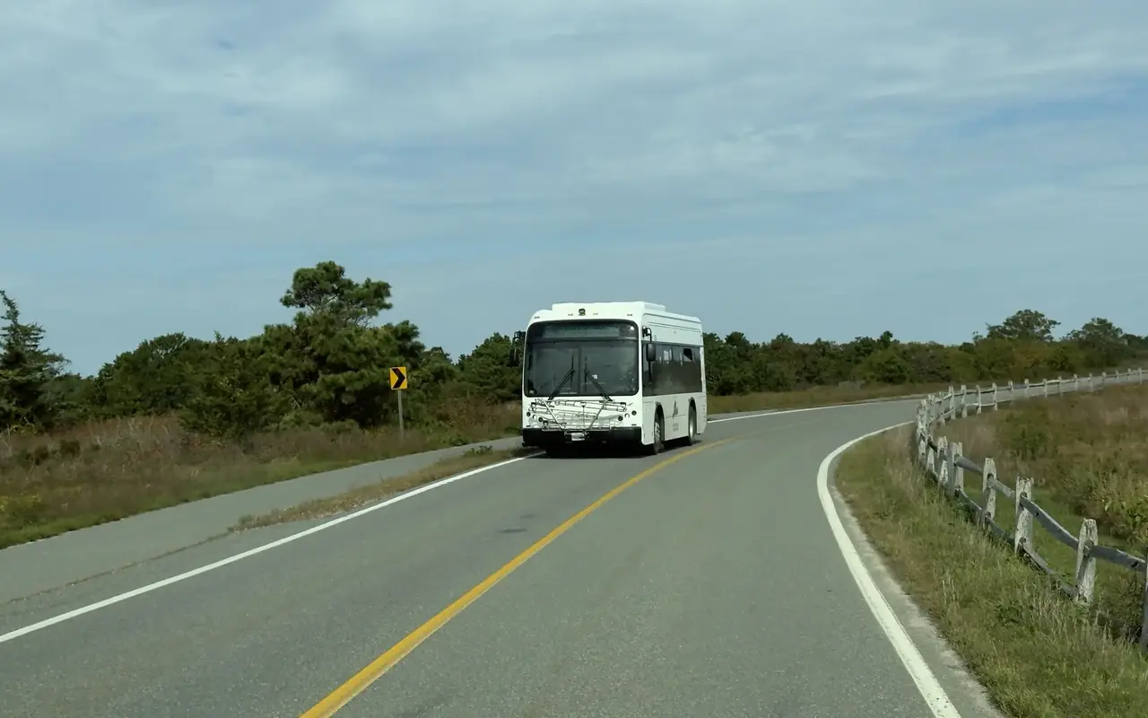 A white VTA bus with a bike rack on the front coming around a curve on the road near South Beach in Katama, Edgartown, a split-rail fence along the right side, scrub oak and pitch pine behind and a paved path alongside on the left, under a high grey-blue sky