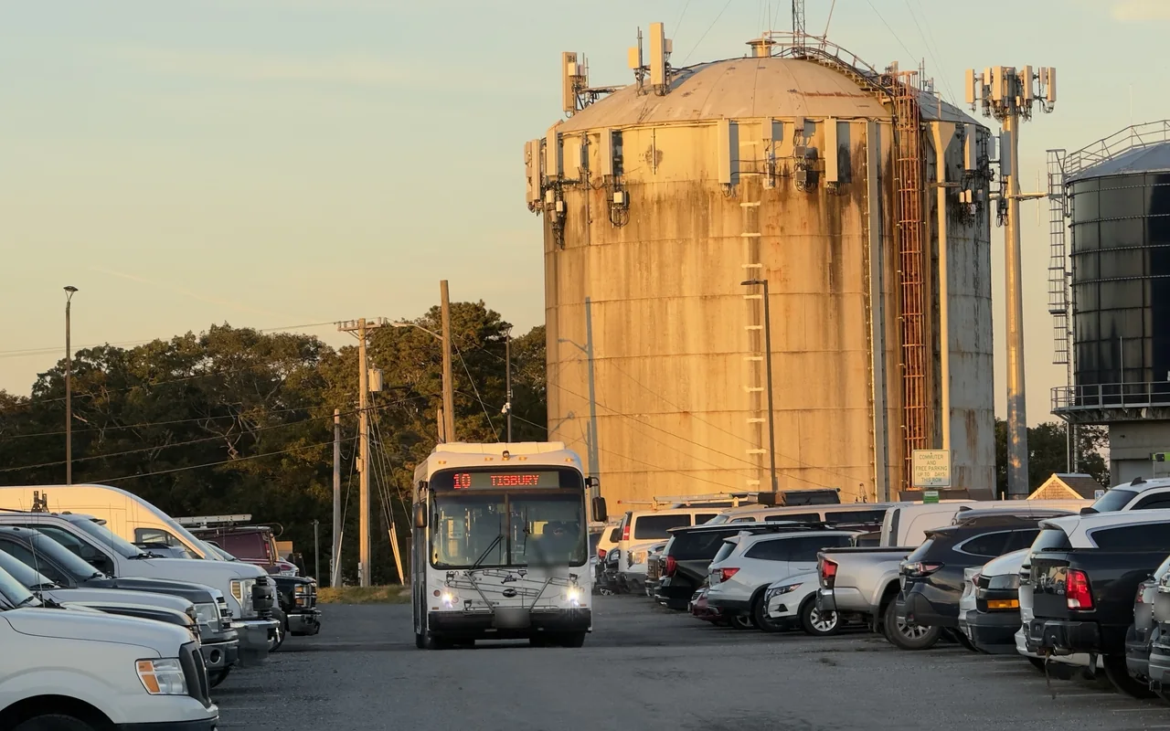 A white VTA bus showing route 10 Tisbury driving between rows of parked cars, two large water tanks behind it in evening light
