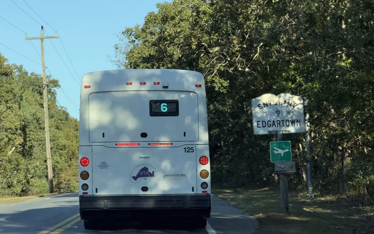 A white VTA bus showing route 6 passing an Entering Edgartown sign with a green airport sign below it on a tree-lined road