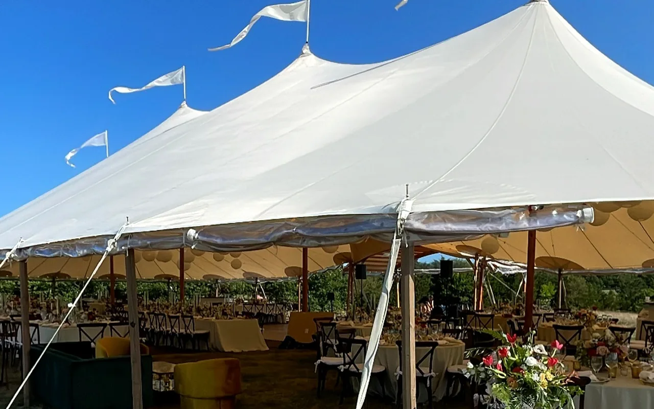 A large white peaked tent with pennant flags on its tops under a blue sky, round dining tables, chairs and paper lanterns inside, a table with flowers in front on dry grass