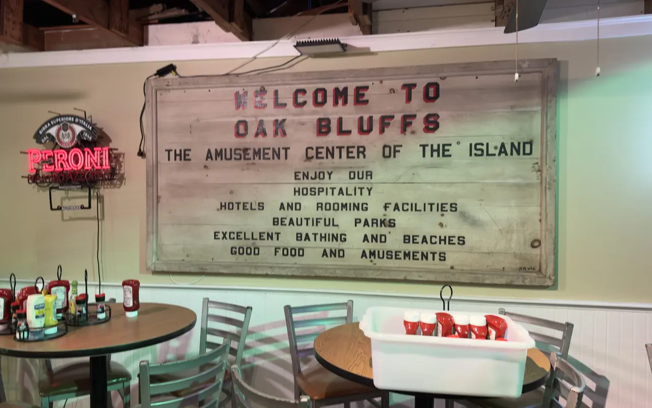 An old painted wooden sign reading Welcome to Oak Bluffs, the Amusement Center of the Island, hanging on the wall inside the Sand Bar and Grille
