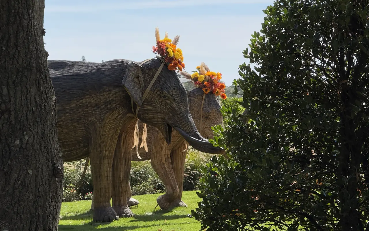 Two life-size woven wicker elephant sculptures with crowns of orange and yellow flowers and pampas grass, standing on a green lawn between a tree trunk and a hedge