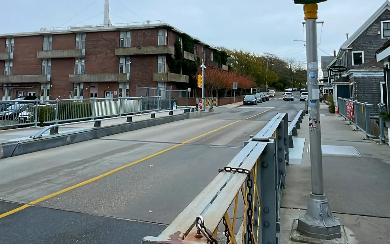 Small steel drawbridge over the Eel Pond channel in Woods Hole with a brick building and village houses beyond