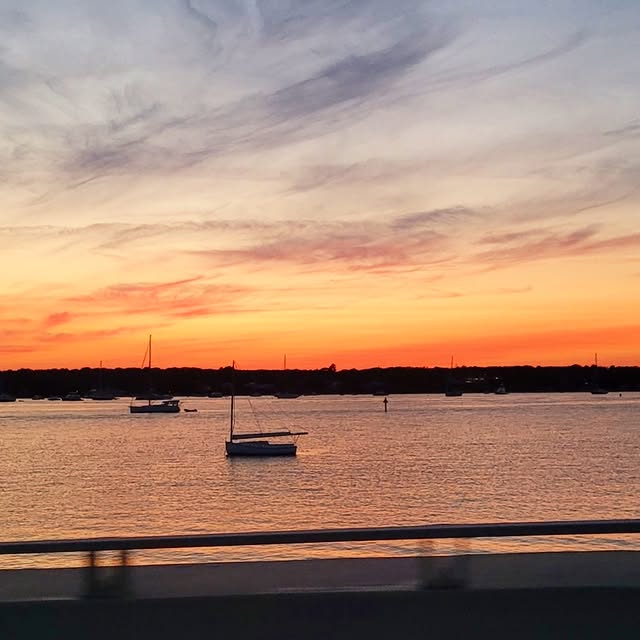 Sunset over Vineyard Haven harbor. Boats on their moorings, one sail still out catching the last of...